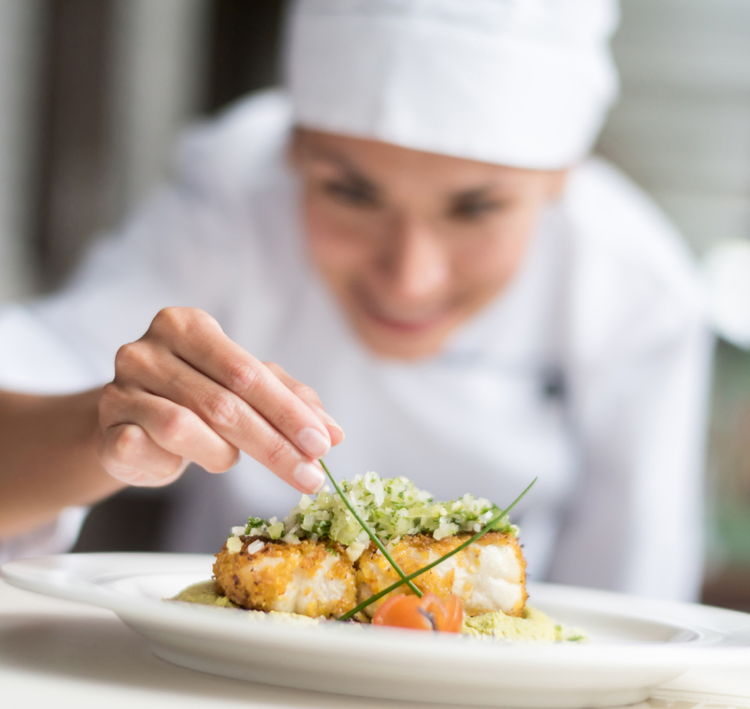 A chef in a white uniform and hat carefully garnishes a plated dish with herbs, focusing intently as if during a cooking class. The dish features breaded fish, diced vegetables, and chives.