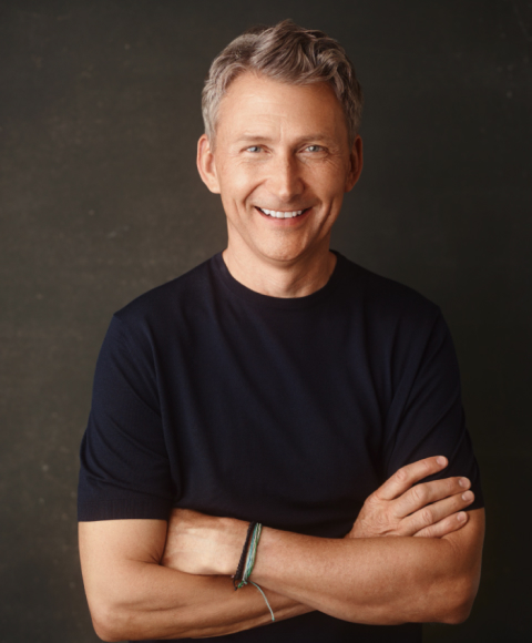 A middle-aged man with short gray hair smiles at the camera, standing against a dark background with his arms crossed, ready to lead engaging sessions while wearing a black t-shirt and bracelets on his left wrist.