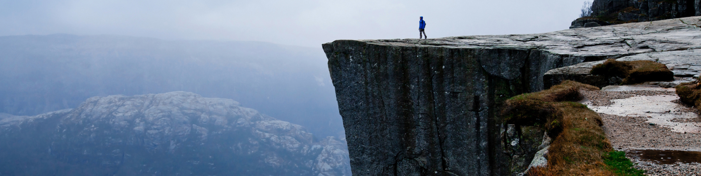 A person in a blue jacket stands on the edge of a steep, rocky cliff overlooking a misty mountain landscape, as if ready to embark on a new course under the cloudy sky.
