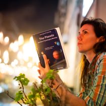 A woman in a plaid shirt sits outdoors at night, holding a book titled The Planets. Warm streetlights and blurred city buildings glow in the background as she gazes thoughtfully into the distance.