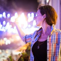 A woman wearing a plaid shirt stands on a balcony at night, reaching her hand out toward blue and white festive lights glowing in the background. She looks to the side, illuminated by the city lights.