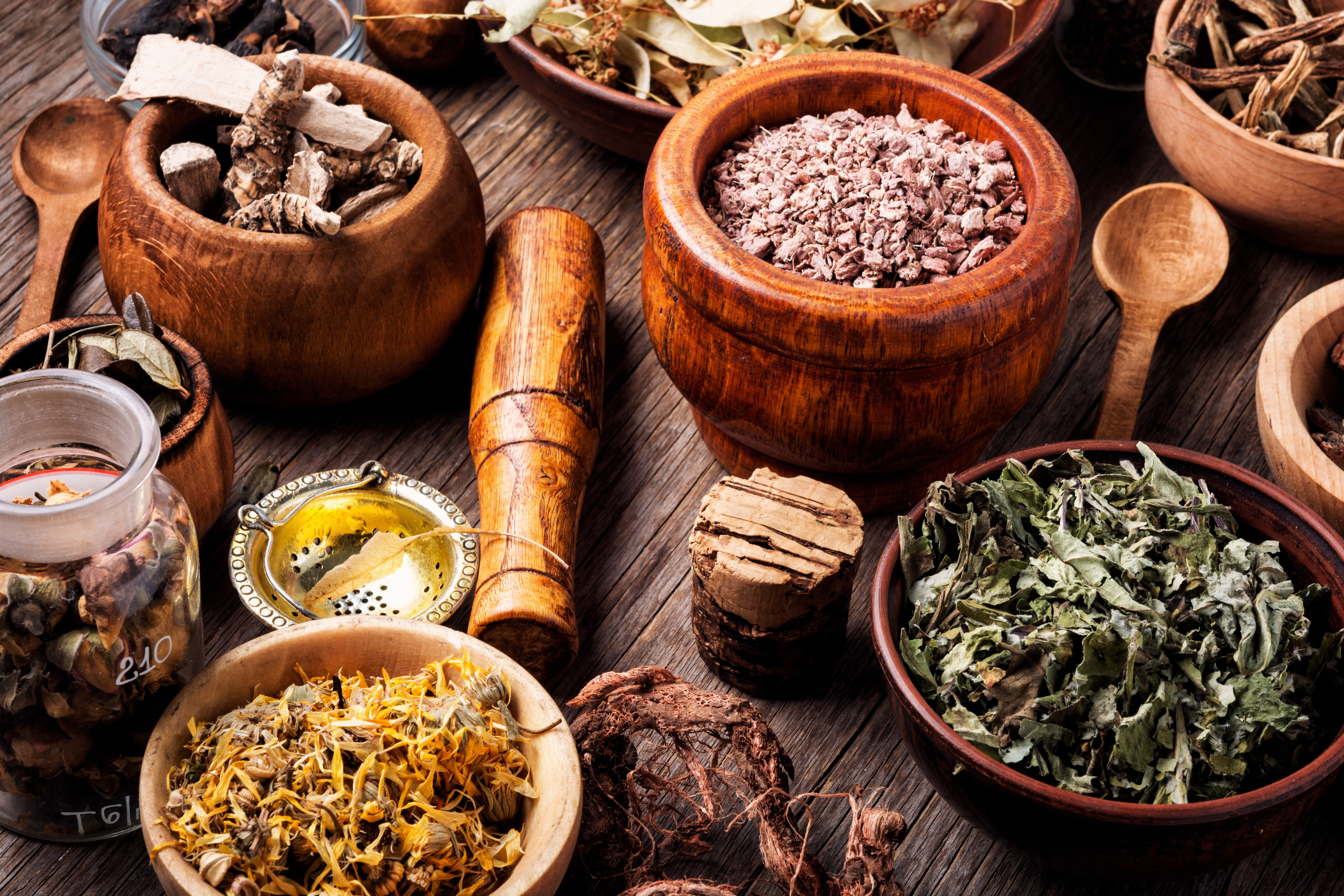 A Plant Medicine Session kit featuring dried herbs, flowers in bowls and jars, a small oil jar, and a wooden pestle is arranged on a rustic table, evoking the feel of a traditional apothecary or herbal medicine setting.