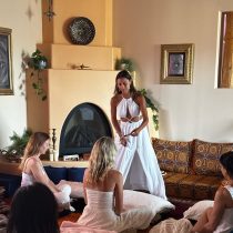 Alexandra is standing in a white dress leading an energy healing program with multiple other women sitting on the ground cross-legged.