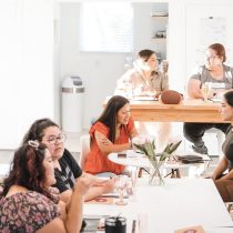 Six women are in a sunlit, modern room. Four sit at a table with flowers, smiling and discussing The Queen Business Coaching Program, while two others chat at the kitchen counter.