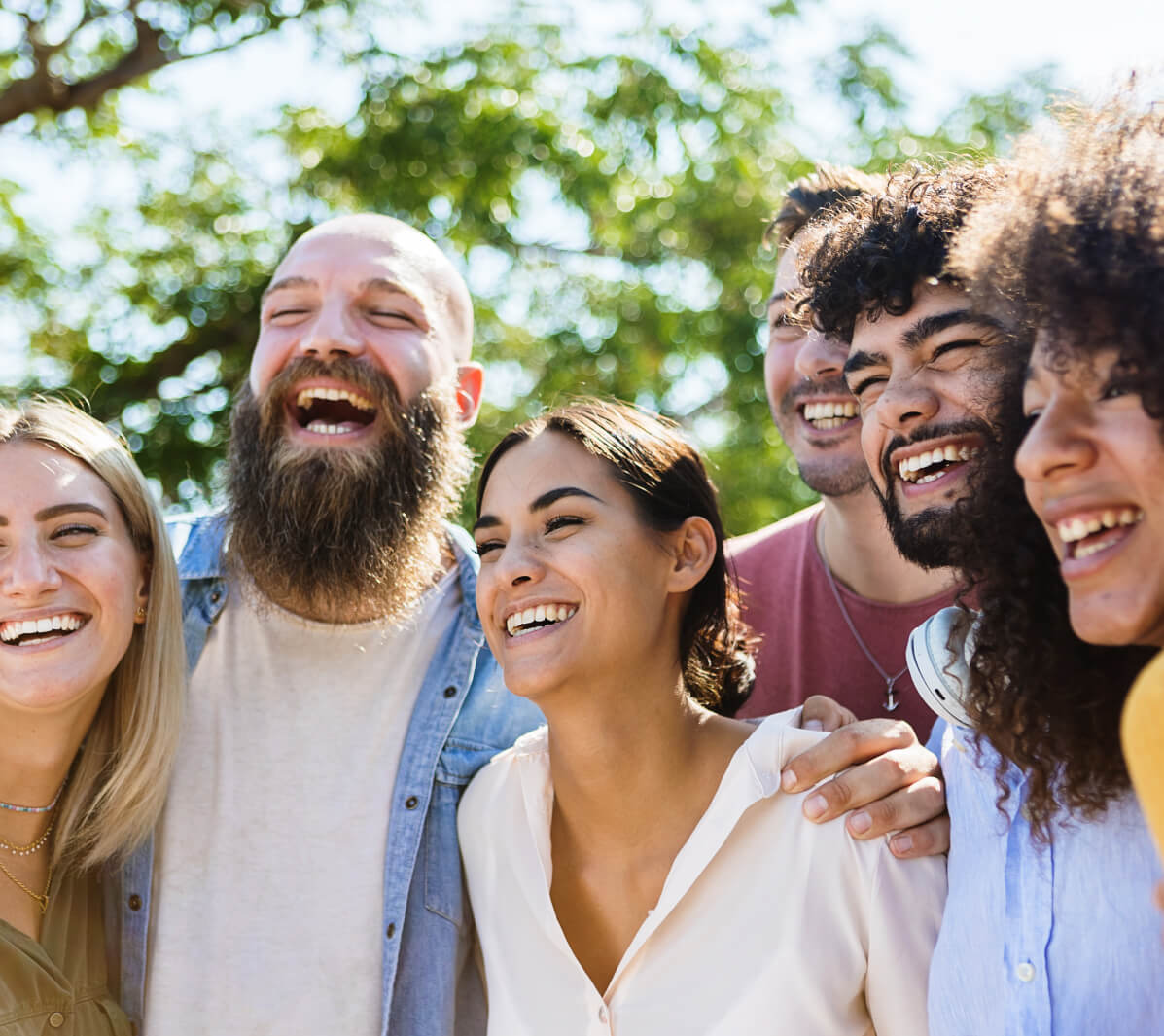 A diverse group of six young adults stand close together outdoors, smiling and laughing under the sunlight, sharing a moment after their coaching sessions, with green trees in the background.