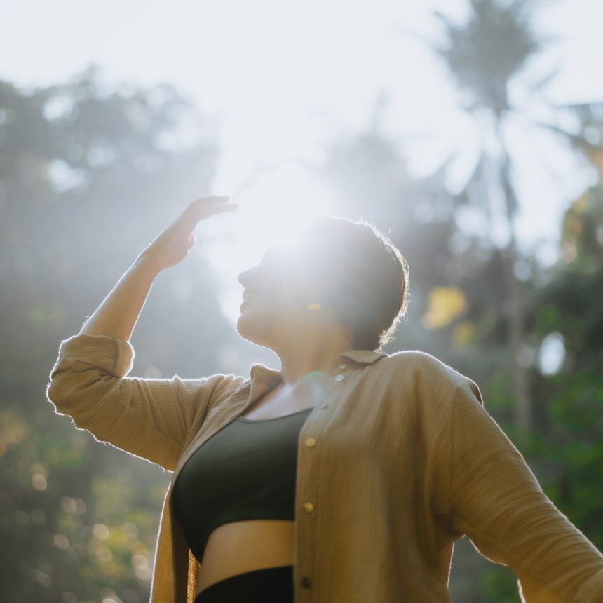 Kristy, whos helps you in finding your purpose, is wearing a black top and bottom set with a long-sleeve tan shirt over it. She is standing in the jungle. The sun is causing a glare in the image. Her hand is lifted up towards her head.