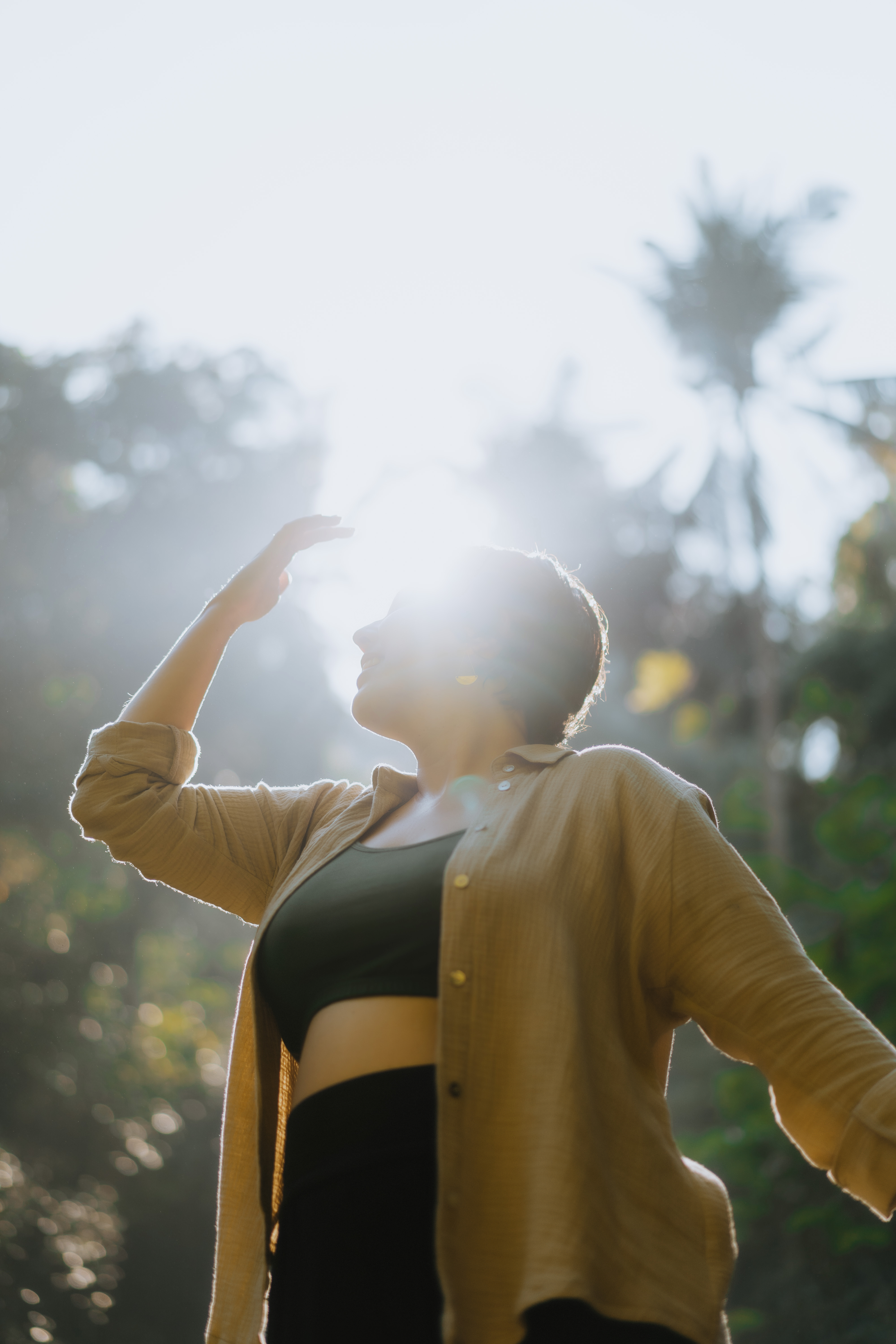 Kristy, whos helps you in finding your purpose, is wearing a black top and bottom set with a long-sleeve tan shirt over it. She is standing in the jungle. The sun is causing a glare in the image. Her hand is lifted up towards her head.
