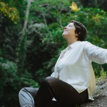 A person sits peacefully outdoors on a rock, arms outstretched and eyes closed, surrounded by sunlight and lush trees—evoking the serenity experienced during a Family Constellations Individual Session.