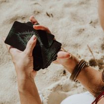 A person sits on sandy ground, holding and shuffling a deck of black cards with green geometric patterns. They wear bracelets on one wrist and a white garment. The scene is sunlit and serene.