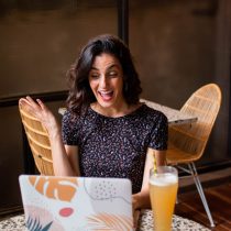 A woman with shoulder-length dark hair smiles and waves at her laptop during a video call, possibly seeking guidance or discussing courses, while sitting at a cafe table with a tall glass of juice beside her.