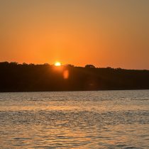 An orange sunset glows over calm water, the sun partially hidden by a tree-lined horizon with gentle ripples—capturing the tranquil essence of Intuitive Healing.
