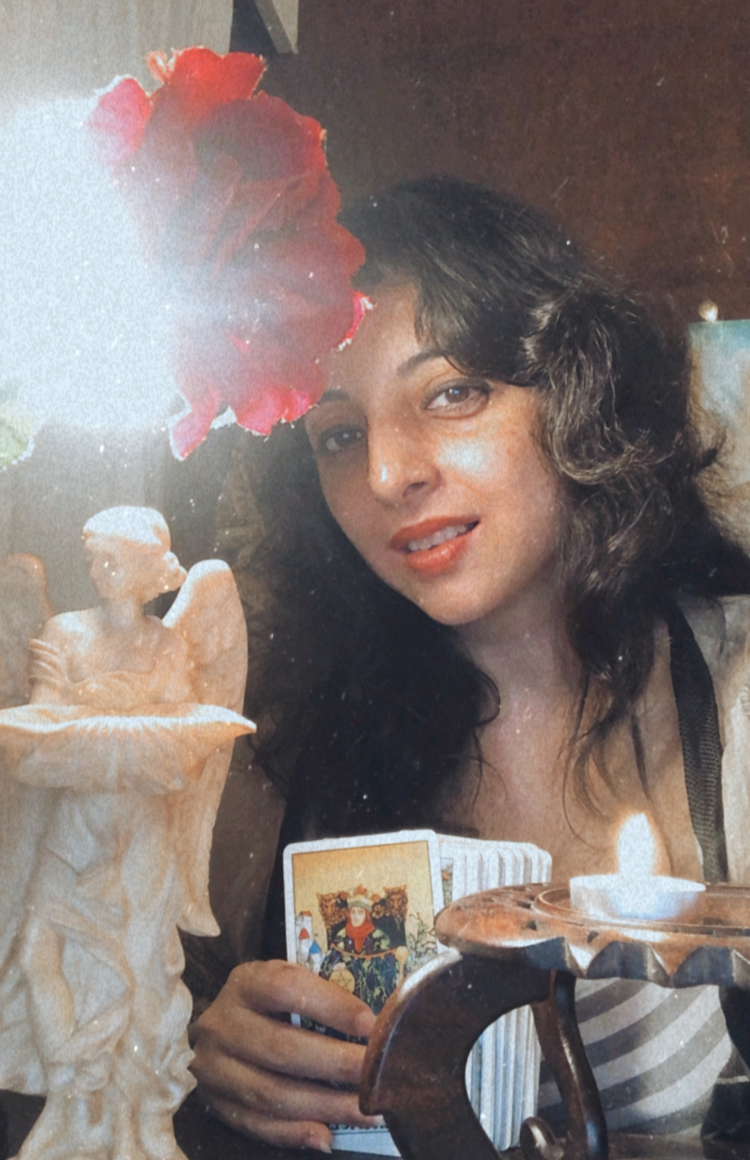 A woman with wavy hair smiles while holding tarot cards. A lit candle, an angel statue, and a red flower are also visible in the warm, softly lit scene.