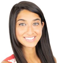 A young woman with long straight dark hair, green eyes, and light brown skin smiles at the camera in a coral top with white lace, reflecting the glow from the 3-Month Mind + Body Reset personalized nutrition coaching against a white background.