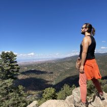A man with a beard and sunglasses stands on a rocky cliff, wearing a black tank top, orange sweatshirt tied around his waist, and pink pants, looking out over a vast valley with trees and mountains under a clear blue sky.