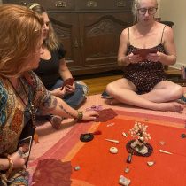 Three women gather for an Intuitive Healing session, sitting around a red cloth adorned with crystals and stones. One reads from a card while the others listen, engaging in a group ritual or spiritual gathering.