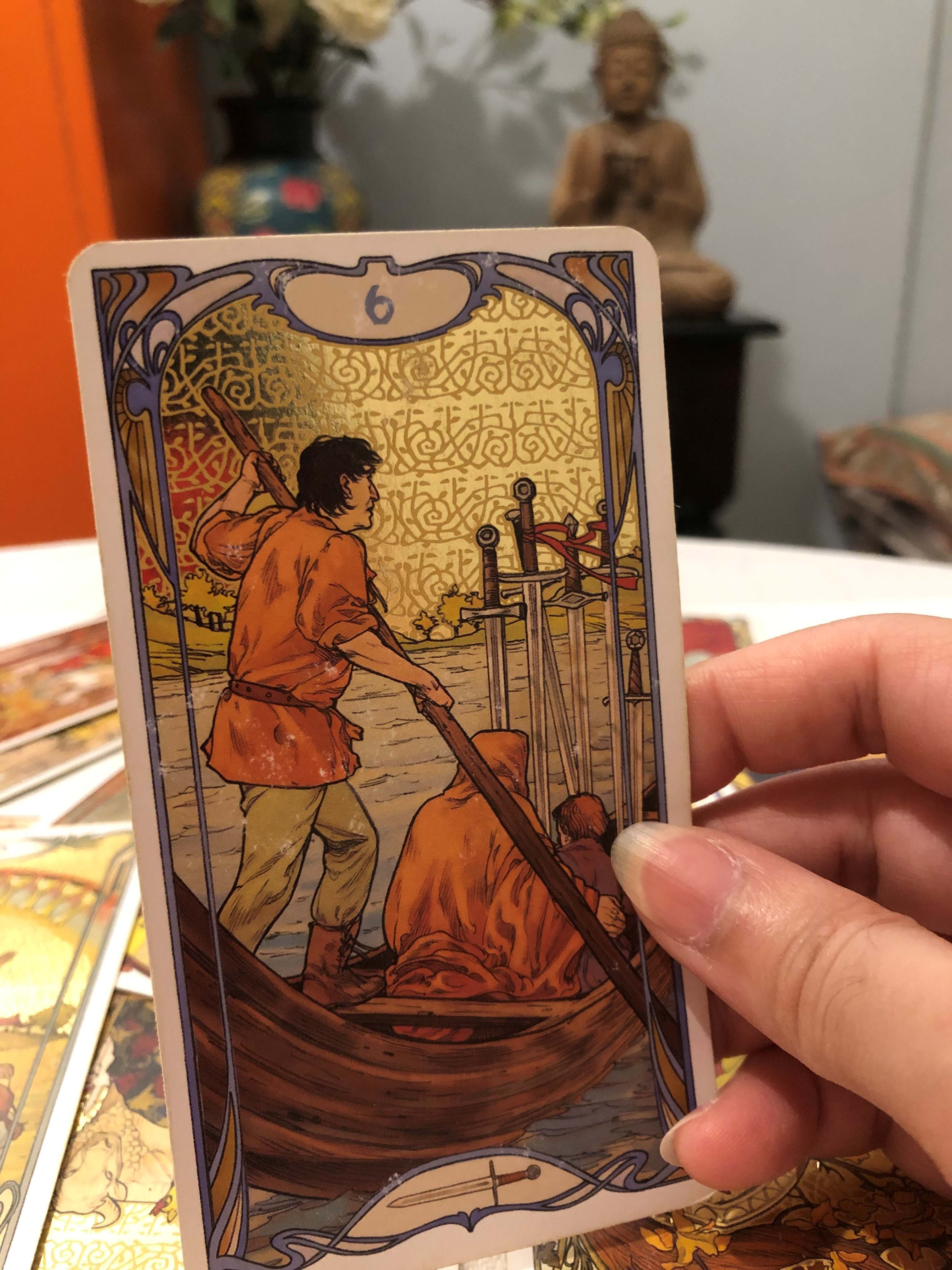 A hand holds a tarot card of a boat with three people and six swords, while other tarot cards are spread on the table. The blurred background features a Buddha statue. Product: Tarot Reading & Spiritual Guidance.