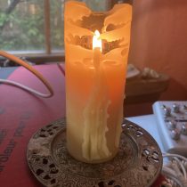 A lit yellow candle with melted wax drips stands on an ornate metal holder, placed beside a red box and in front of a window with greenery outside.