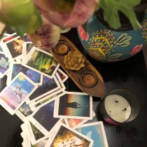 A scattered Tarot Reading and Meditation deck lies on a dark table beside a black candle, a decorative vase with flowers, and a wooden holder with two tea lights and a yellow flower.