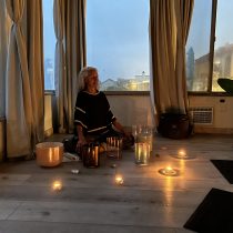 Roxy is wearing a black shirt and is sitting crossed leg in the corner of a room with her eyes closed. She is surrounded by her sound bath meditation bowls.