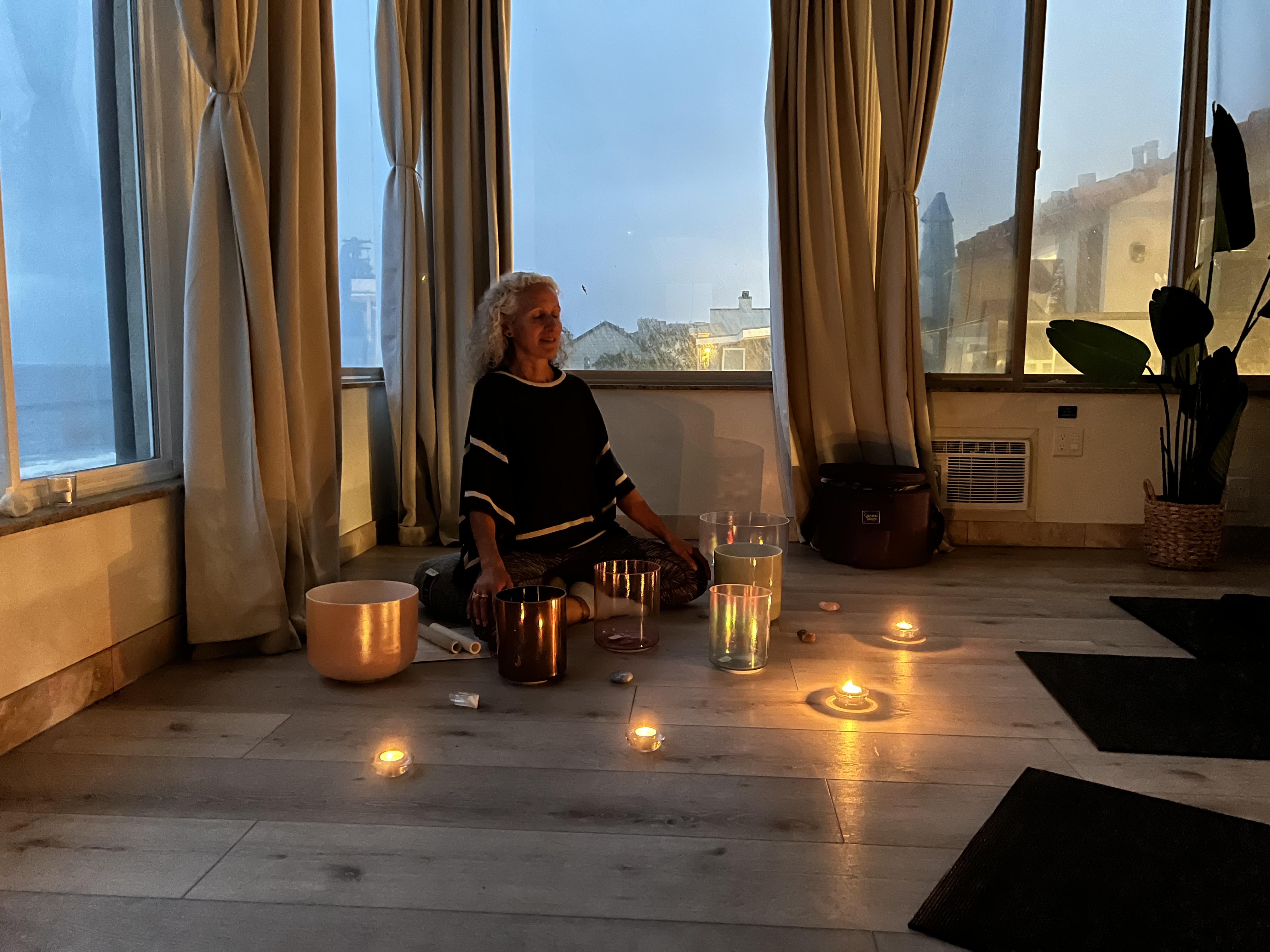 Roxy is wearing a black shirt and is sitting crossed leg in the corner of a room with her eyes closed. She is surrounded by her sound bath meditation bowls.