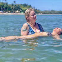 Two women are in the ocean near a tropical beach. One, with a tattoo, supports another floating calmly; both appear relaxed and serene. Palm trees and blue sky set the scene for their peaceful moment with Intuitive Healing.