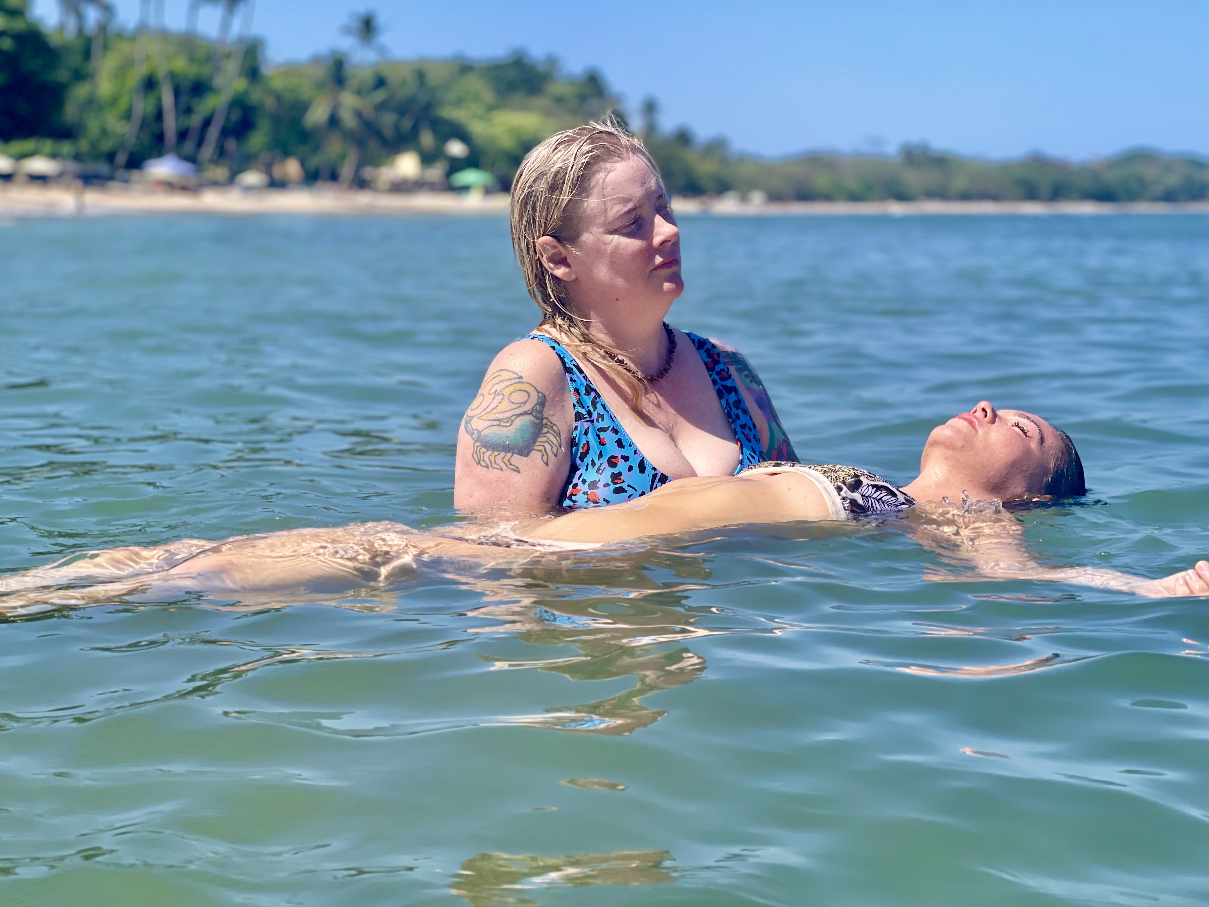Two women are in the ocean near a tropical beach. One, with a tattoo, supports another floating calmly; both appear relaxed and serene. Palm trees and blue sky set the scene for their peaceful moment with Intuitive Healing.