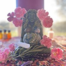 The Intuitive Healing statue features a smiling woman with long hair surrounded by pink flowers, a globe, stones, crystals, and greenery on a colorful cloth in soft natural light.