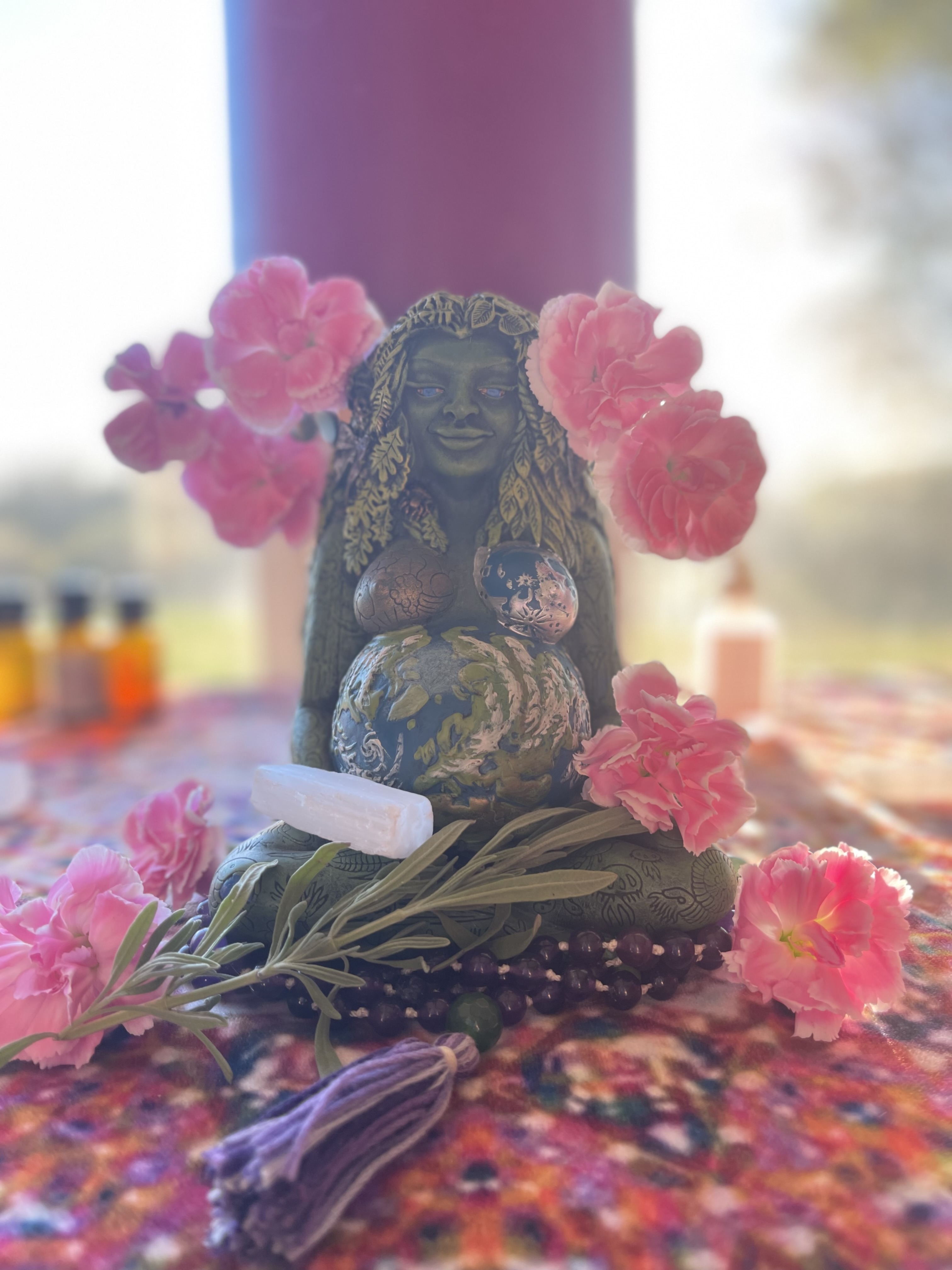 The Intuitive Healing statue features a smiling woman with long hair surrounded by pink flowers, a globe, stones, crystals, and greenery on a colorful cloth in soft natural light.