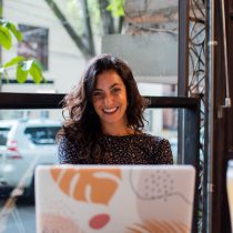 A woman with curly hair smiles while sitting at a table in front of a window, using her laptop with a colorful abstract cover—ready for online coaching sessions. Outside, trees and parked cars can be seen.