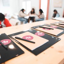 Notebooks with pens are on a wooden table in the foreground, while a group discusses The Queen Business Coaching Program at a blurred table in the bright, airy room.
