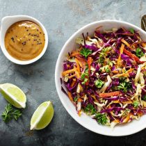An overhead view of an Asian style salad with purple cabbage, red bell peppers, orange carrots, and green cilantro. There is a peanut sauce in a ramekin next to it with two slices of lime. This is a recipe from one of Julia's personalized nutrition coaching program meal plans.