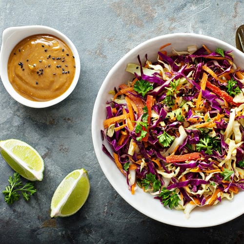 An overhead view of an Asian style salad with purple cabbage, red bell peppers, orange carrots, and green cilantro. There is a peanut sauce in a ramekin next to it with two slices of lime. This is a recipe from one of Julia's personalized nutrition coaching program meal plans.