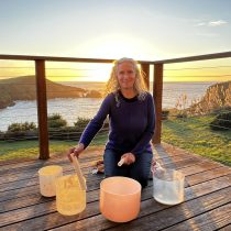 At sunset on a wooden deck, a woman with curly hair plays crystal singing bowls as part of the Intuitive Sound Therapy Coaching Package of 3. The ocean and cliffs glow in the golden background, enhancing the serene atmosphere.