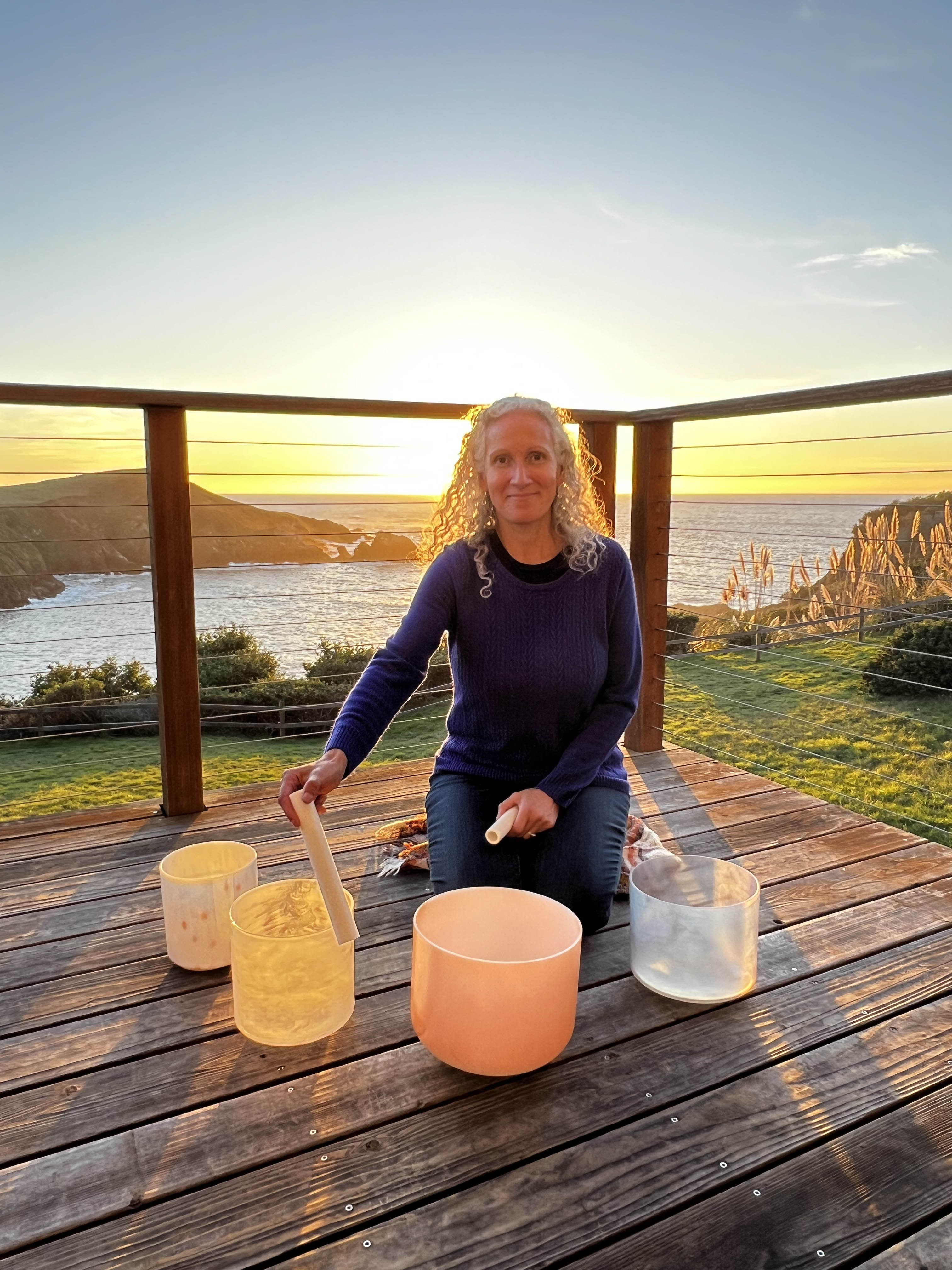 At sunset on a wooden deck, a woman with curly hair plays crystal singing bowls as part of the Intuitive Sound Therapy Coaching Package of 3. The ocean and cliffs glow in the golden background, enhancing the serene atmosphere.