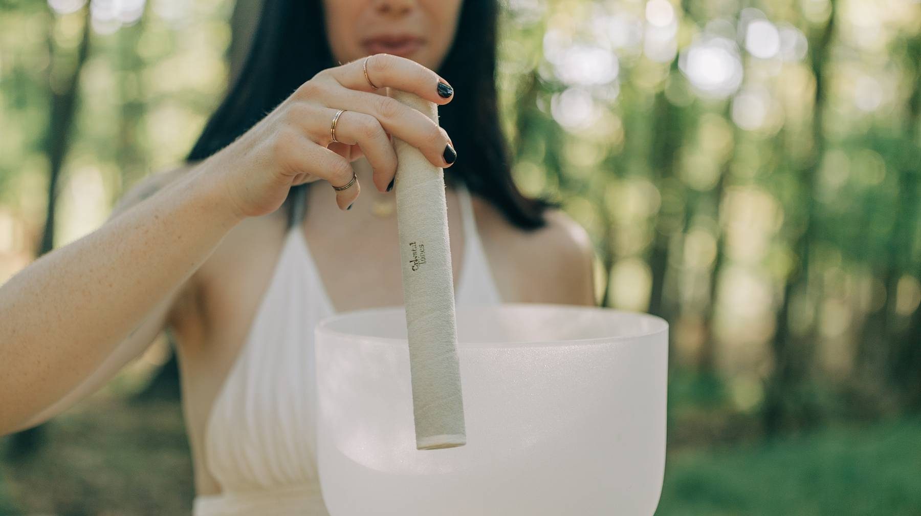 A woman in a white top raises a mallet above a frosted crystal singing bowl outdoors, capturing the peaceful essence of the Sound Healing & Intuitive Guidance Session against a softly blurred forest background.
