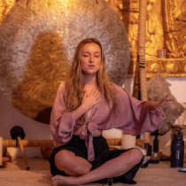 A woman sits cross-legged on the floor with one hand on her chest and the other extended outward, eyes closed, in front of a large gong. Blue and white roses and ritual objects are arranged on the floor beside her.
