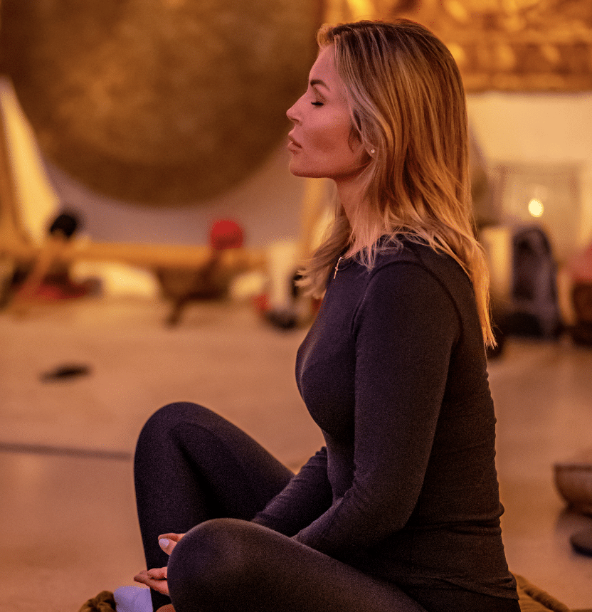 A woman with blonde hair sits cross-legged indoors, eyes closed, meditating in a warm, softly lit room with golden and brown decor in the background.