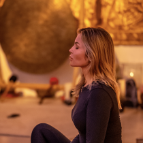 A woman with blonde hair sits cross-legged indoors, eyes closed, meditating in a warm, softly lit room with golden and brown decor in the background.