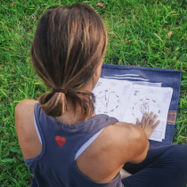 A person with brown hair in a ponytail, wearing a sleeveless shirt with a red heart on the back, sits on grass reviewing Love Compatibility Reading charts.