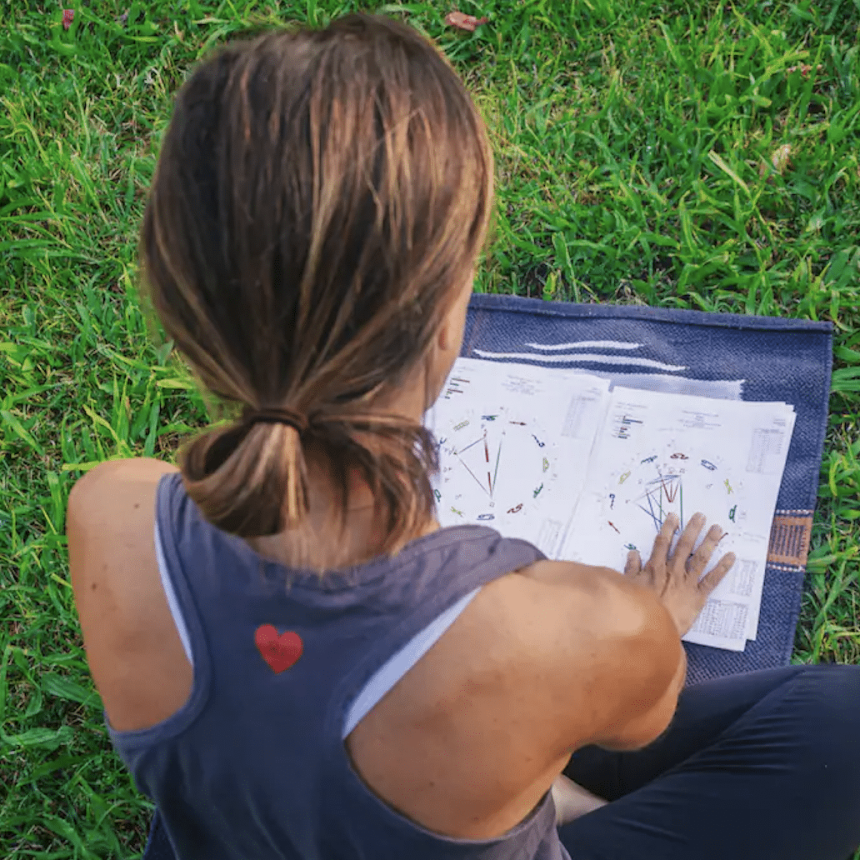 A person with brown hair in a ponytail, wearing a sleeveless shirt with a red heart on the back, sits on grass reviewing Love Compatibility Reading charts.