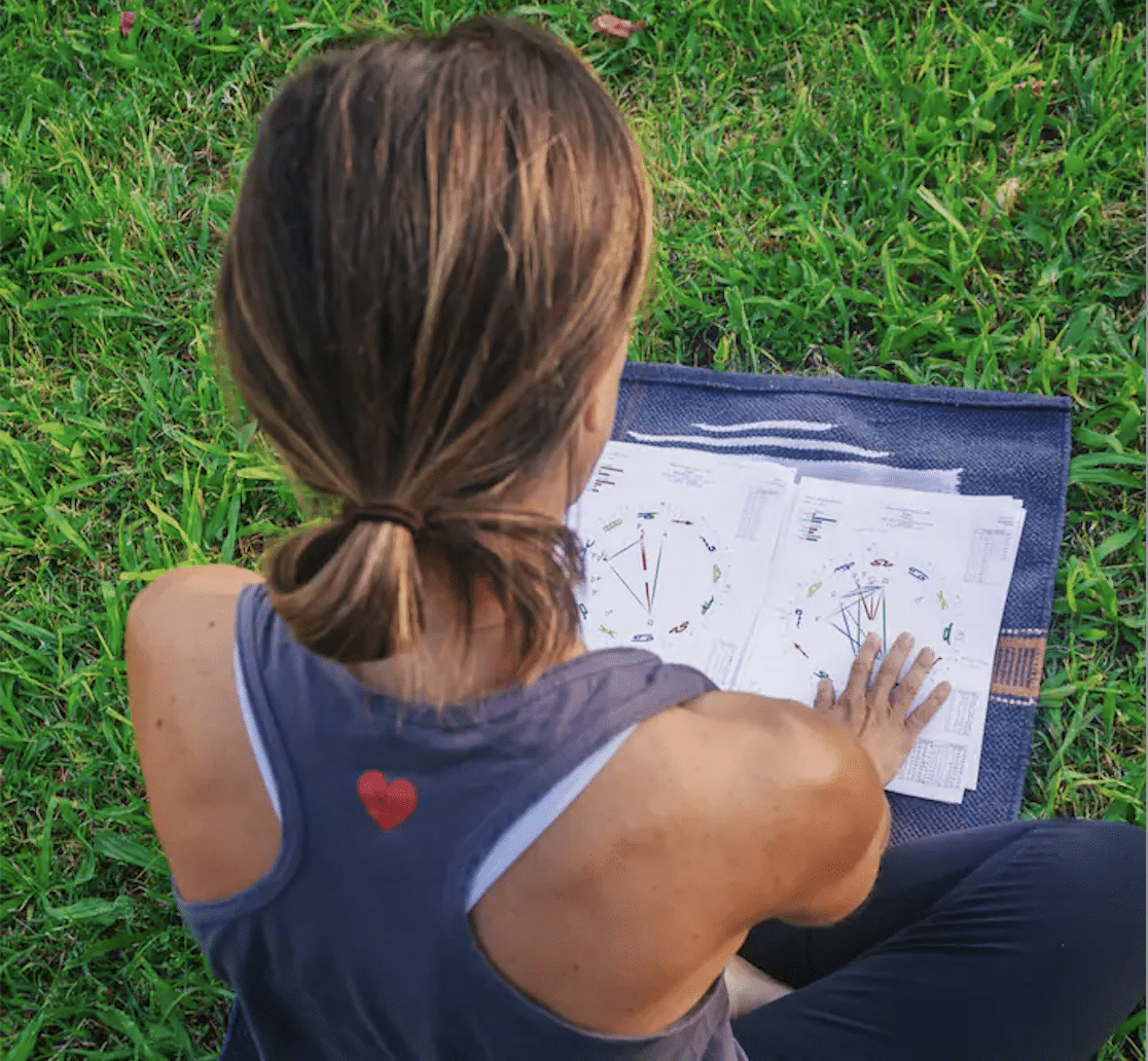 A person with brown hair in a ponytail, wearing a sleeveless shirt with a red heart on the back, sits on grass reviewing Love Compatibility Reading charts.