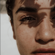 A close-up of a face in natural light, with leaf-shaped shadows cast across the skin, captures only one eye, part of the nose, and lips—highlighting texture and detail. Inspired by Breakthrough.