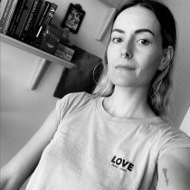 Black and white selfie of a person with long hair, wearing a LOVE t-shirt and hoop earrings, standing before shelves with books, art, and materials for Unleash Your Potential online personal coaching. The person has a neutral expression.