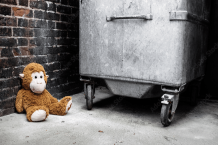 A brown stuffed monkey sits alone on the ground next to a large metal dumpster in an alleyway with brick walls, embodying what it feels like when youre feeling lost in life.