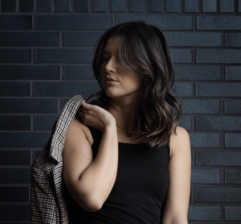 A woman with shoulder-length dark hair, wearing a black sleeveless top and holding a plaid jacket over her shoulder, stands in front of a dark brick wall, embodying the spirit of the You vs You Journey: 3 months product. Her face is partly hidden by her hair.