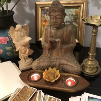 A tabletop for Intuitive Therapy, Tarot, Life Navigation features a Buddha statue, angel figurine, framed quote, tarot cards, notebook, two tealight candles in a lotus holder, and a vase with flowers.