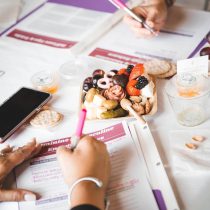 Two people work on worksheets near a phone, candle, crackers, and a tray of fruit, cheese, berries, and snacks—setting the scene for The Queen Business Coaching Program’s casual workshop with light refreshments.