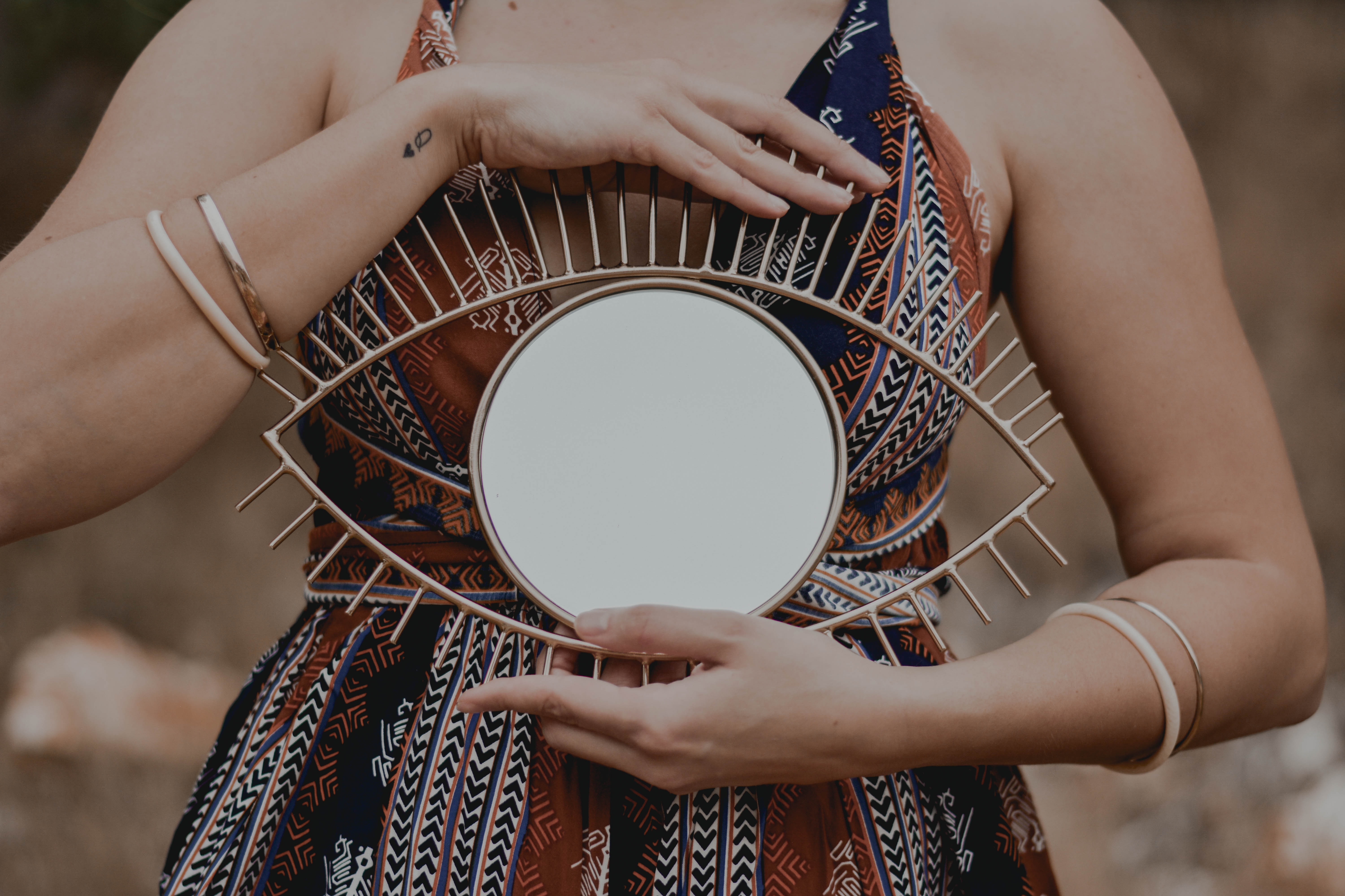 A person in a patterned dress holds an eye-shaped mirror with gold lashes at chest height, reflecting the spirit of our Intuitive Readings & Psychic Guidance. Their bracelet-adorned arms and torso are visible, creating a mystical ambiance.