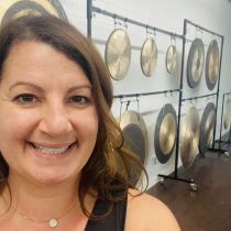 A smiling woman takes a selfie in front of several large gongs hanging on metal stands in a room with white walls.