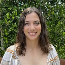A woman with wavy brown hair and a light striped top smiles at the camera while standing outdoors in front of lush green foliage.